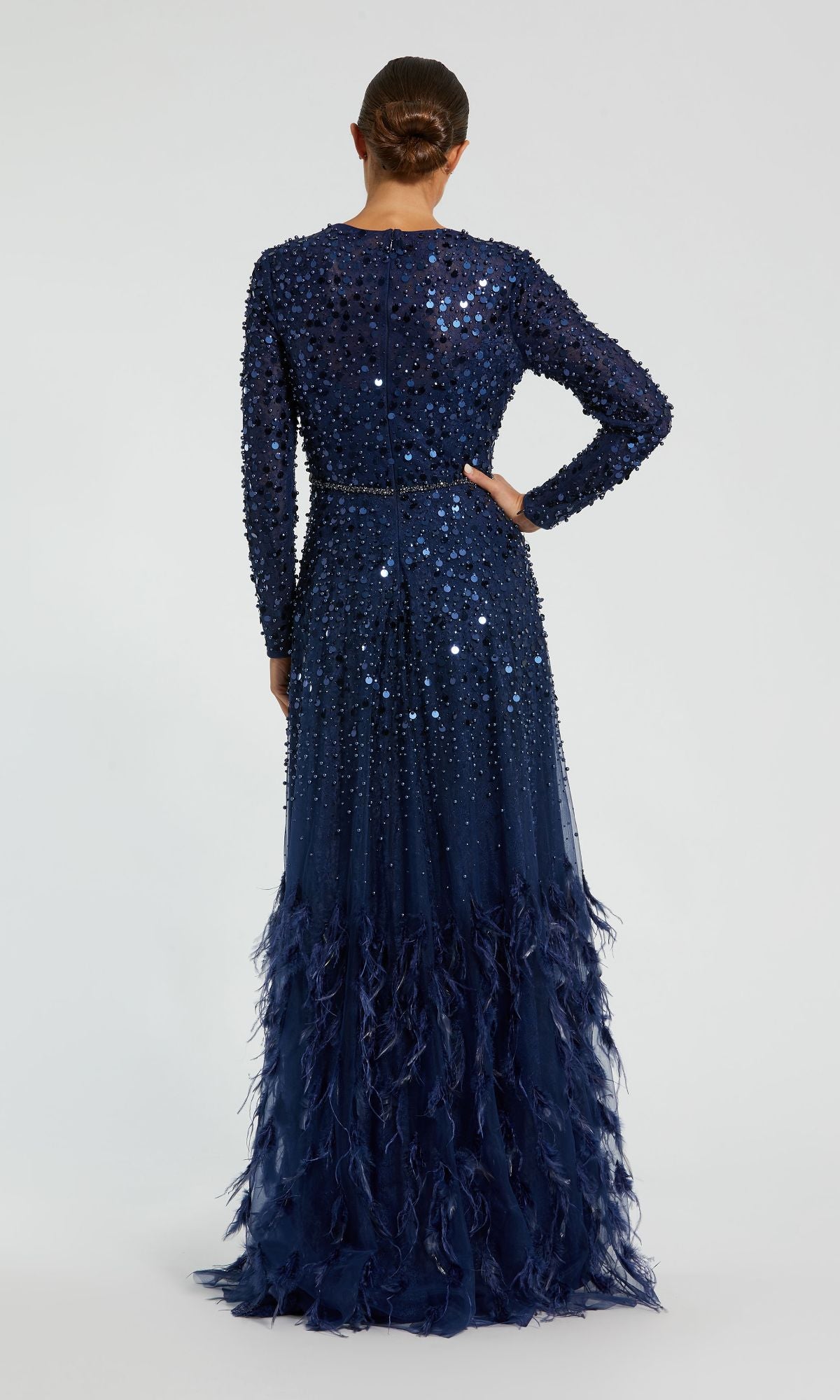A woman faces away, wearing the Mac Duggal 11782 Long Sleeve Sequin Formal Gown with floor-length sequins and feathered hem. Her hair is in a low bun—an elegant choice for a mother of the bride dress.
